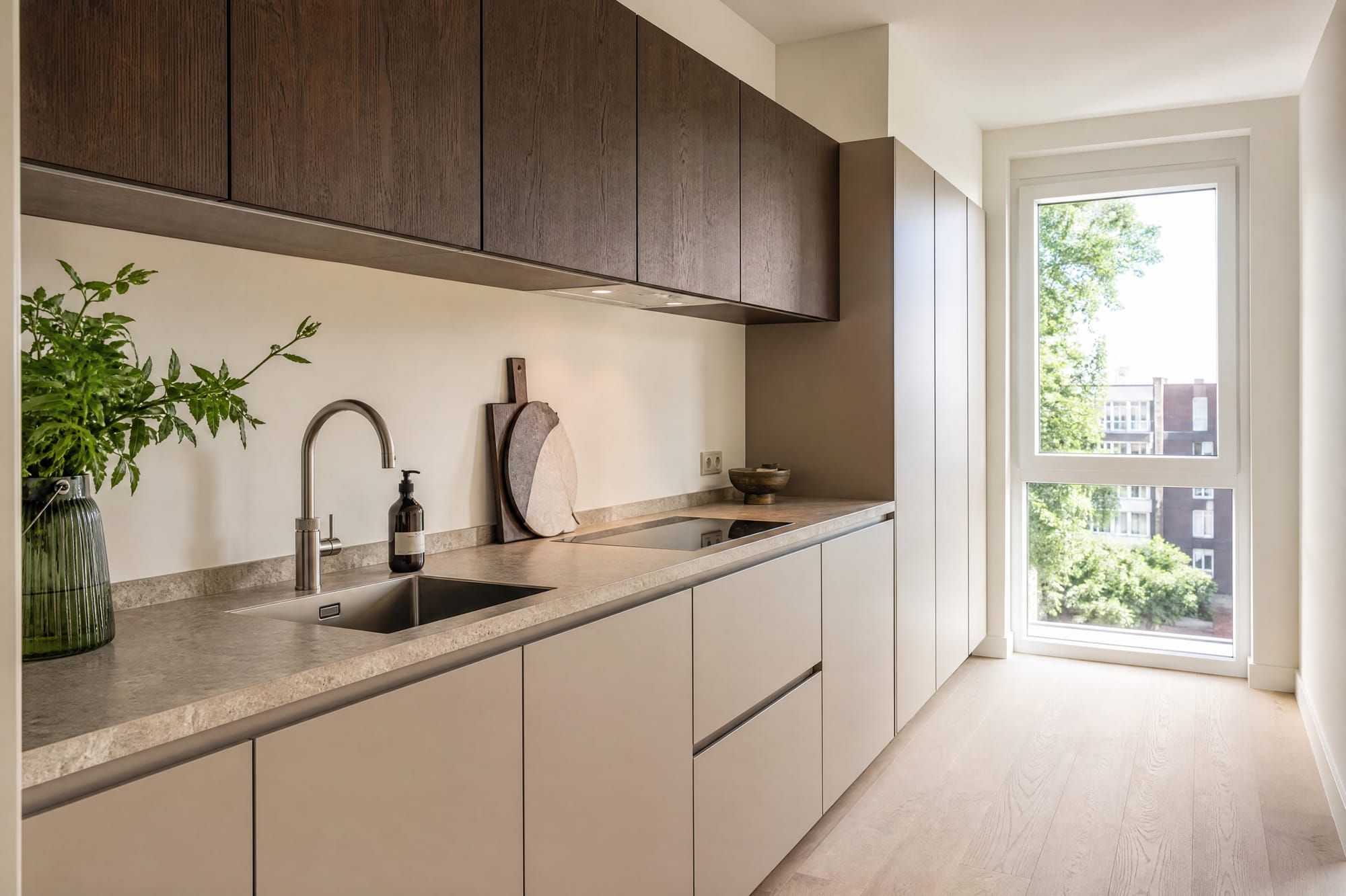 Strakke greige keuken met donker-eikenhouten bovenkasten en hoog raam.