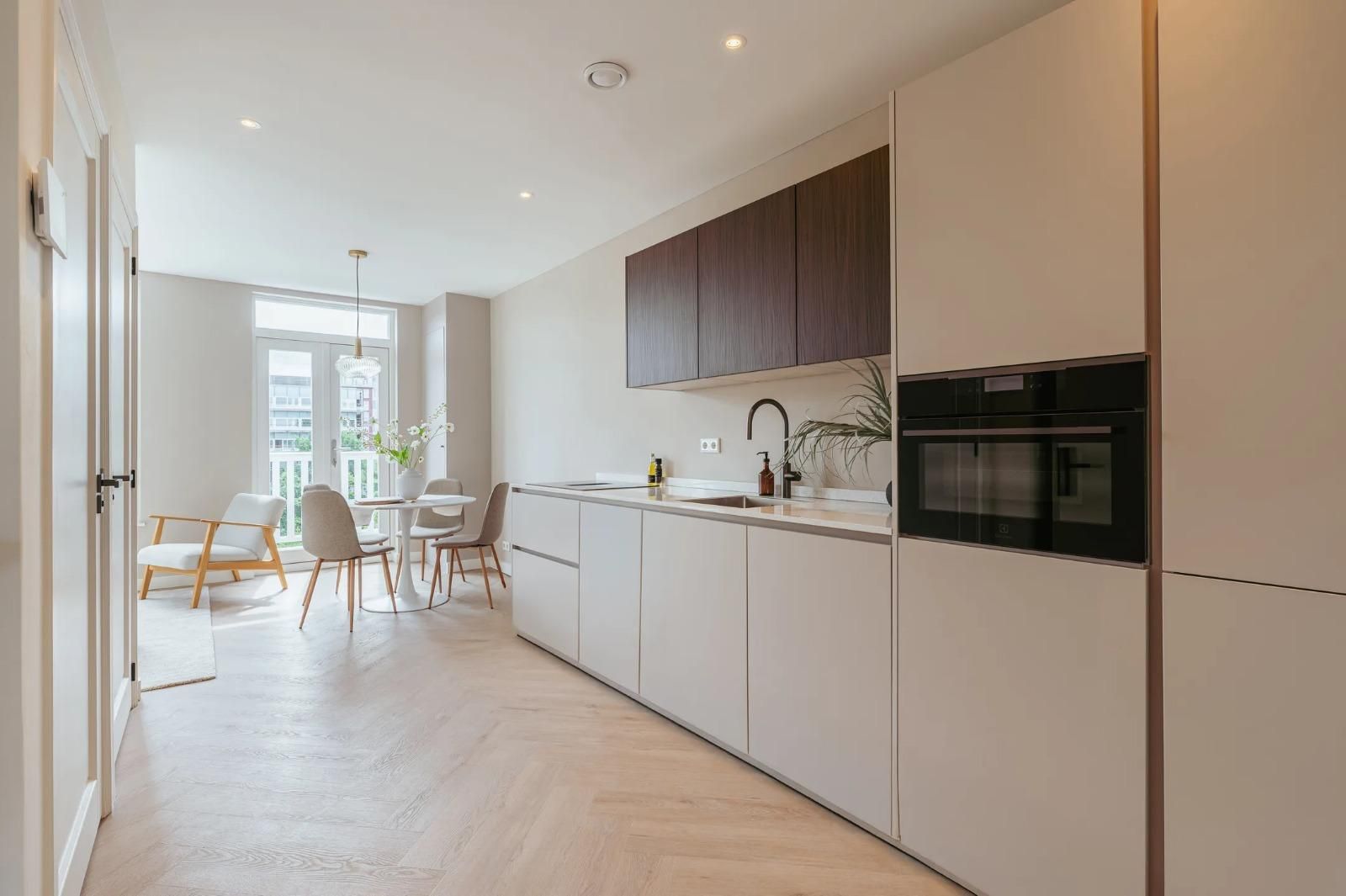 Lichte keuken met crème fronten, houten bovenkasten en eethoek bij het balkon.